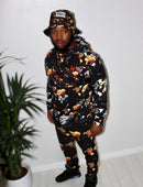 Man wearing Ember Blaze Men’s Hoodie with vibrant print and matching bucket hat, standing in a modern setting.