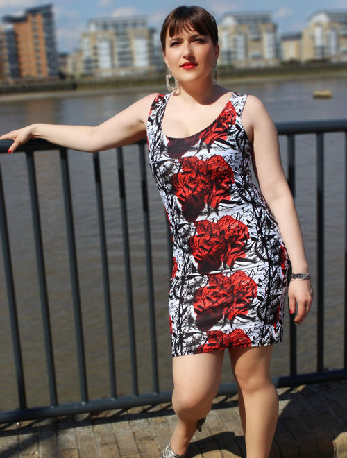 Woman posing in Red Rose Bodycon dress, showcasing vibrant floral print against a modern backdrop by the water.