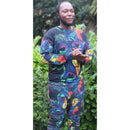 Man wearing a colorful geometric peacock design sweater and pants, standing outdoors with greenery in the background.